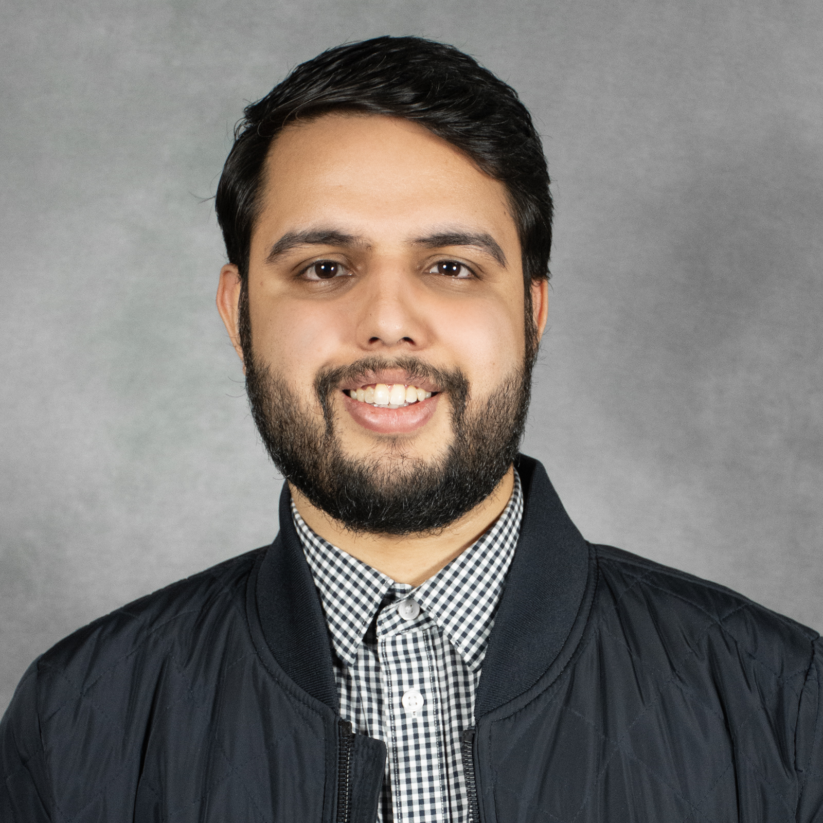 Man with short black hair and beard wearing checked shirt and black jacket