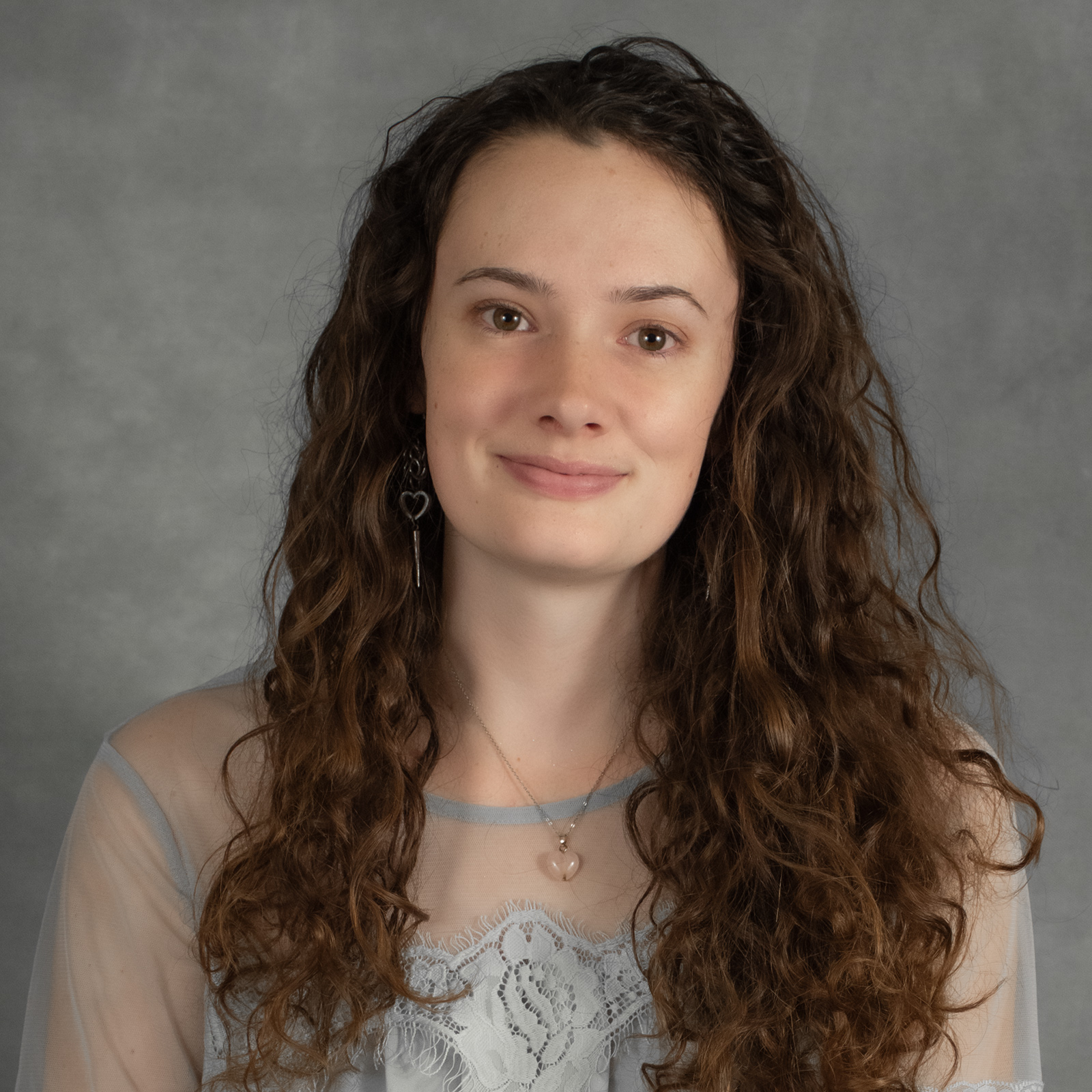 Woman with long brown wavy hair wearing a light gray shear top with lace