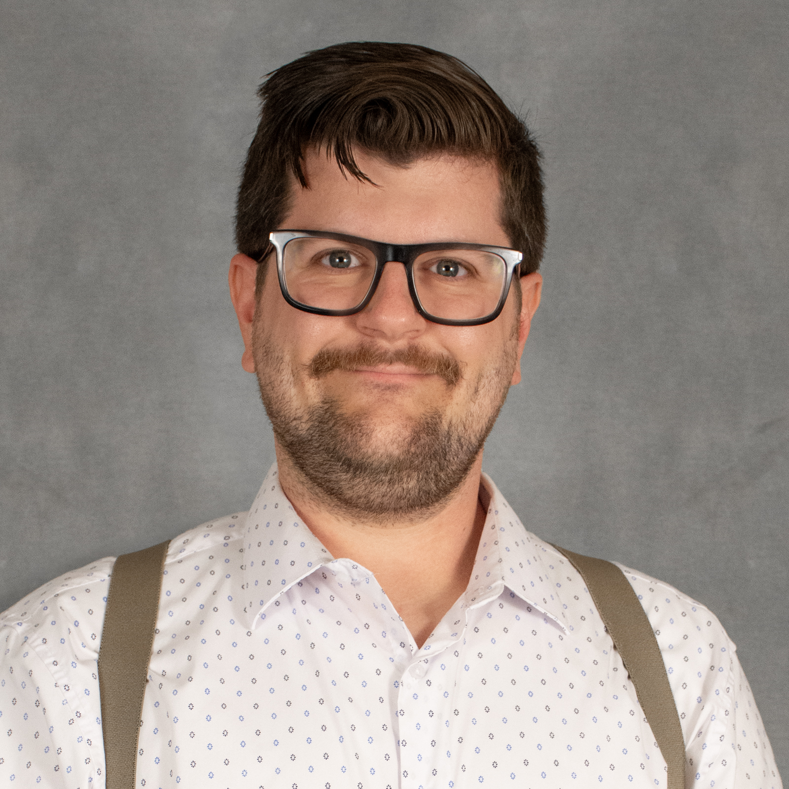 Man with ark brown short hair, short beard wearing glasses, white shirt and suspenders