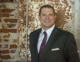 white man in a suit standing in front of a brick wall smiling