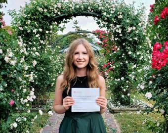 young white woman in a green dress holding a certificate in a garden surrounded by flowers