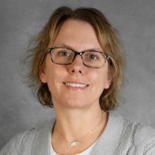 Woman with short brown air wearing a gray sweater and gold necklace