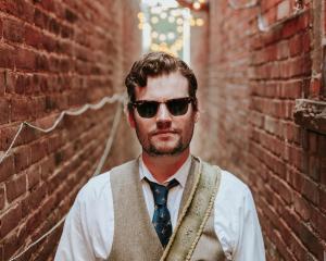 white man in sunglasses wearing a white fhirt and tan best standing in between two brick walls