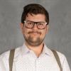 Man with ark brown short hair, short beard wearing glasses, white shirt and suspenders